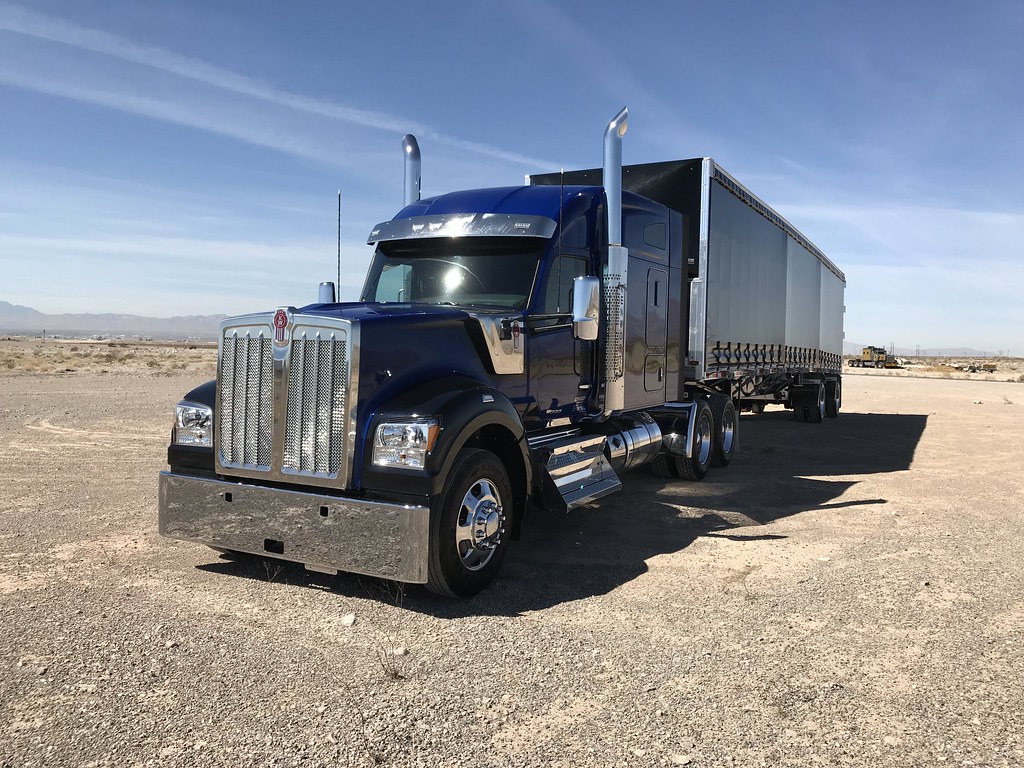 Blue semi truck with chrome grille and long trailer parked on a dry desert landscape under a clear blue sky.